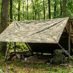 Camouflaged tarnplanen tarp stretched as a forest shelter between trees, showing a realistic outdoor survival setup with ropes and natural woodland background.