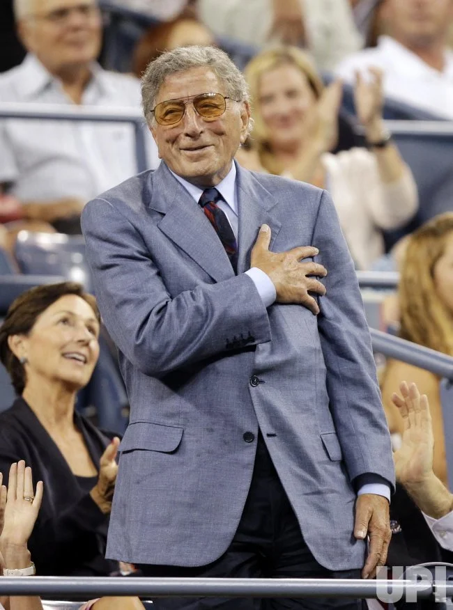 Tony Bennett attends a New York sports event, standing among spectators in a blue blazer with his hand on his chest while the crowd applauds and celebrates his appearance.