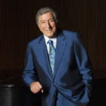 Tony Bennett NewYork portrait featuring the legendary singer in a blue suit, smiling beside a grand piano against a dark elegant backdrop, reflecting his timeless music legacy and connection to New York.
