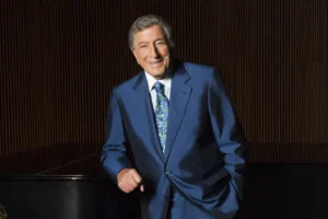 Tony Bennett NewYork portrait featuring the legendary singer in a blue suit, smiling beside a grand piano against a dark elegant backdrop, reflecting his timeless music legacy and connection to New York.