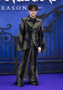 Isaac Ordonez ethnicity article image showing the young actor wearing a stylish black and olive striped suit while posing on a blue carpet event backdrop with decorative gate elements.
