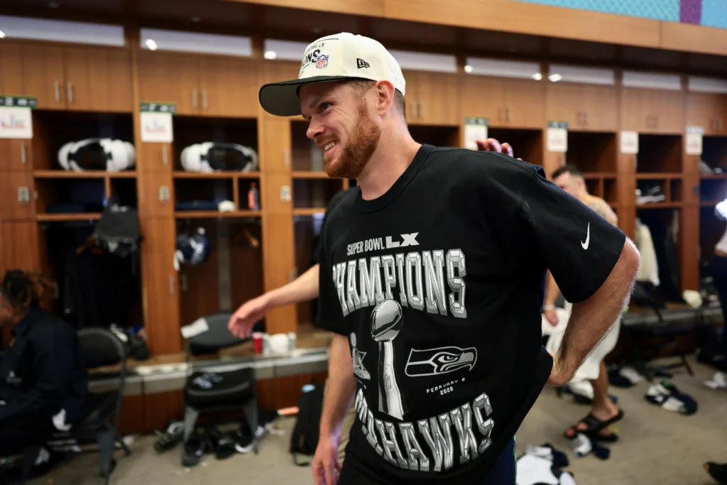 Sam Darnold draft class quarterback Sam Darnold standing in a team locker room wearing a Super Bowl Champions T-shirt and cap, smiling as he celebrates a championship victory with teammates in the background.