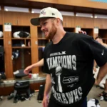 Sam Darnold draft class quarterback Sam Darnold standing in a team locker room wearing a Super Bowl Champions T-shirt and cap, smiling as he celebrates a championship victory with teammates in the background.