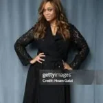 **SEO Optimized Alt Text with Keyword:** **Tyra Banks NewYork appearance at a formal event, wearing a black long-sleeve dress and posing confidently against a blue-gray backdrop.**
