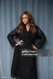 **SEO Optimized Alt Text with Keyword:** **Tyra Banks NewYork appearance at a formal event, wearing a black long-sleeve dress and posing confidently against a blue-gray backdrop.**