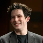 **Josh O'Connor ears visible as the actor smiles during a public appearance in a dark blazer and black shirt**