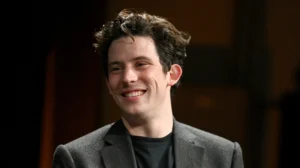 **Josh O'Connor ears visible as the actor smiles during a public appearance in a dark blazer and black shirt**
