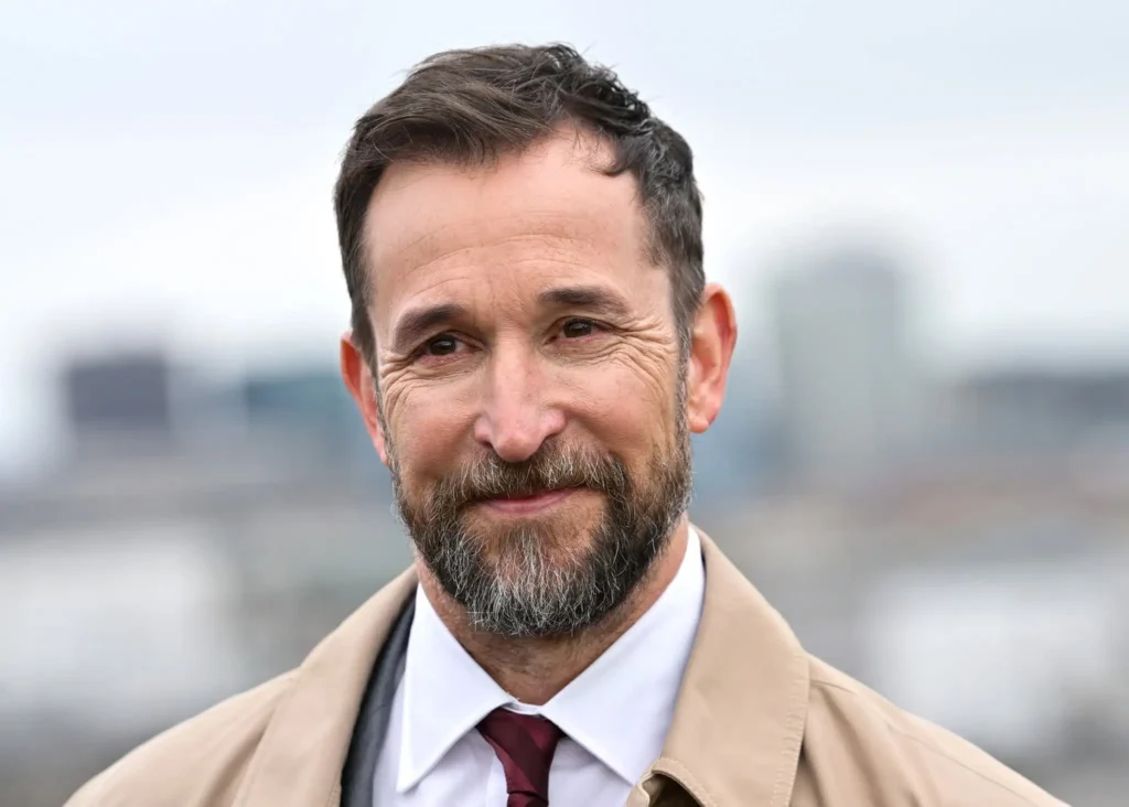 Noah Wyle ER star Noah Wyle smiling in a close-up outdoor portrait, wearing a beige coat, white shirt, and burgundy tie with a blurred city skyline in the background.