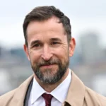 Noah Wyle ER star Noah Wyle smiling in a close-up outdoor portrait, wearing a beige coat, white shirt, and burgundy tie with a blurred city skyline in the background.