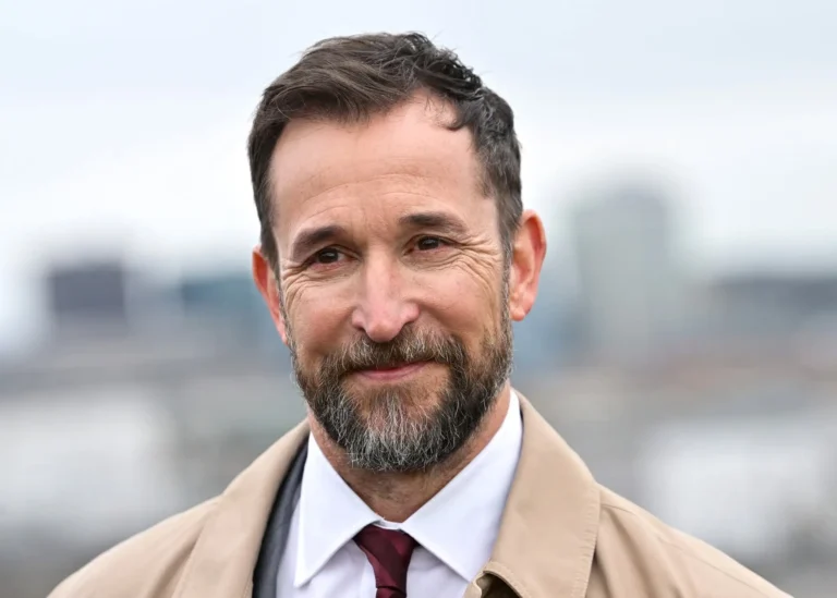 Noah Wyle ER star Noah Wyle smiling in a close-up outdoor portrait, wearing a beige coat, white shirt, and burgundy tie with a blurred city skyline in the background.