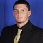 "Professional headshot of a man wearing a black suit, light gold tie, and white dress shirt against a solid blue studio background."