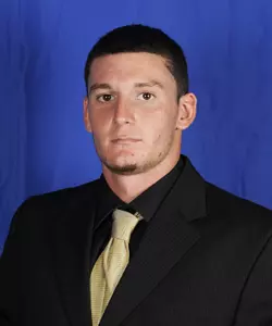 "Professional headshot of a man wearing a black suit, light gold tie, and white dress shirt against a solid blue studio background."
